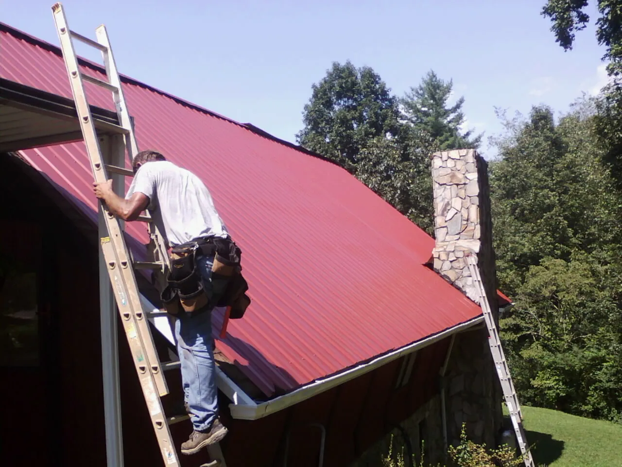 Roofer on a ladder beside a red metal roof and stone chimney — Soffit & Fascia Repair by Roofing Brothers in Pleasant View